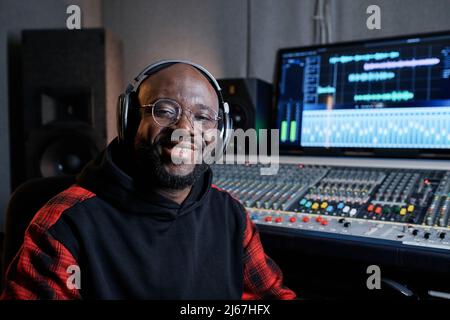 Ritratto di allegro produttore di musica afro-americana indossando cuffie e occhiali seduti in studio di registrazione sorridendo alla macchina fotografica Foto Stock