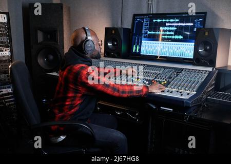 Produttore di registrazione afro-americano professionista che indossa le cuffie sedute alla console di mixaggio lavorando su una traccia musicale Foto Stock