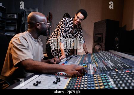 Il produttore di musica afro-americana maturo e il giovane cantante chiacchierano su qualcosa di divertente mentre lavorano nello studio di registrazione Foto Stock