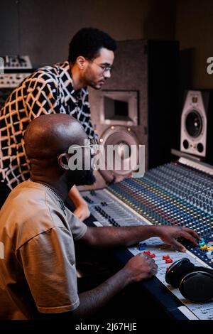 Ripresa verticale di un moderno produttore e giovane cantante afroamericano maturo che crea una traccia musicale in studio di registrazione Foto Stock