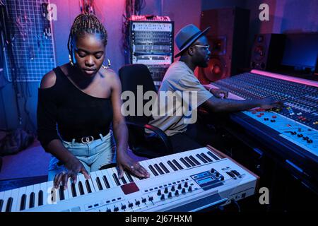 Moderna giovane musicista afroamericana che suona la tastiera digitale mentre tecnico del suono lavora al mixer in studio di registrazione Foto Stock