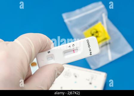 Mano con guanto che contiene una striscia di test dell'antigene SARS-Cov-2 che mostra un risultato negativo. Un tampone nasale all'interno di una sacca è visibile sullo sfondo. Foto Stock