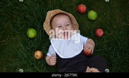 Felice neonato bambino in estate panama cappello cadere giù adagiato su erba a piedi nudi in estate Sunny Day con frutta fresca mele. Bambino bambino bambino ragazzo Foto Stock