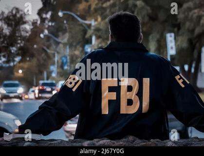 Agente FBI maschile che indossa un cappotto blu scuro con logo FBI che guarda lungo la strada con le auto al tramonto visto da dietro. Foto Stock