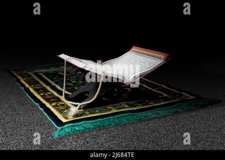 Quran si apre su un placemat di legno con perle di preghiera su un tappeto di preghiera con sfondo nero Foto Stock