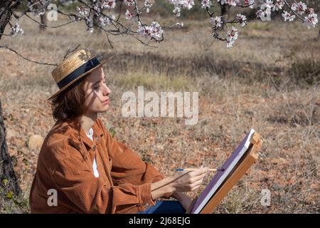 Giovane pittore che dipinge con oli in un campo di mandorli che indossa un cappello Foto Stock