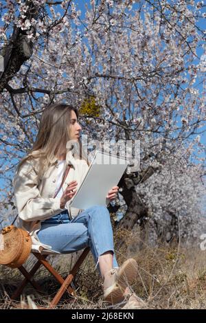 Giovane donna ispirata alla natura mentre dipinge nel suo libro di schizzo circondato dalla natura Foto Stock