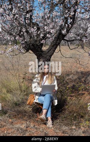 Ritratto di fronte di una giovane e bella donna seduta e appoggiata sul tronco di mandorla in fiore durante il disegno Foto Stock