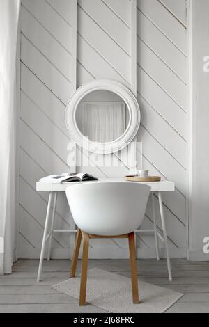 Sedia bianca al tavolo con libro e tazza di bevande poste vicino alla parete con specchio rotondo nella sala luminosa progettata in stile minimal Foto Stock