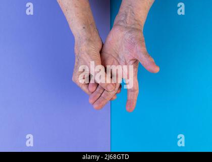 Le mani di una donna anziana sfregano le sue dita irritate su uno sfondo blu e viola. Artrite consapevolezza mese concetto. Spazio di copia. Foto Stock