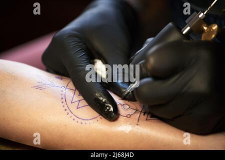 Vista ravvicinata di un tatuaggio irriconoscibile in guanti di gomma che disegnano un tatuaggio con pistola elettrica per tatuaggio Foto Stock