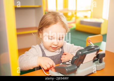 Una bambina di due anni gioca con i giocattoli nella sala giochi. Centro di sviluppo, figlia Kindergarten preme i pulsanti. Registratore di cassa per giocare alla luce del sole del negozio Foto Stock