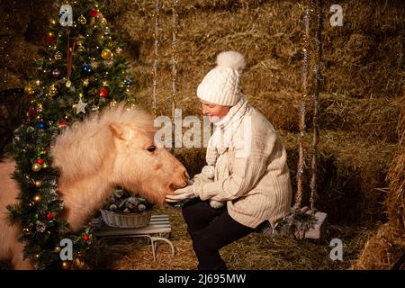 Decorazioni natalizie sulle scuderie. Un bel pony con una corona attorno al collo. Albero di Natale con palloncini, zona foto per il nuovo anno. La donna più anziana si siede sull'altalena Foto Stock