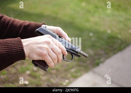 Un giovane uomo carica una pistola contro un'erba verde. Arma da fuoco all'aperto. Sport, hobby primo piano Foto Stock