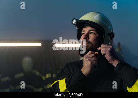 Vigile del fuoco maturo che si prepara all'azione nella stazione dei vigili del fuoco di notte, guardando la macchina fotografica. Foto Stock