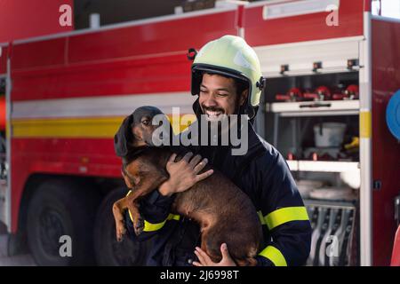 Felice uomo vigile del fuoco che tiene il cane e guarda la macchina fotografica con il camion del fuoco in background Foto Stock