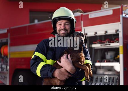 Felice uomo vigile del fuoco che tiene il cane e guarda la macchina fotografica con il camion del fuoco in background Foto Stock