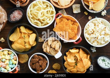 Cibo o spuntini malsani. Tutti i classici spuntini di patate con arachidi, anelli di popcorn e cipolle e pretzel salati in piatti da ciotola su vecchio sfondo di legno. Foto Stock