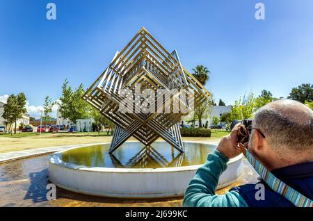 Huelva, Spagna - 28 aprile 2022: Un fotografo che fa una foto al monumento a cubo di acciaio inossidabile nei giardini del Campus de “El Carmen” della H Foto Stock