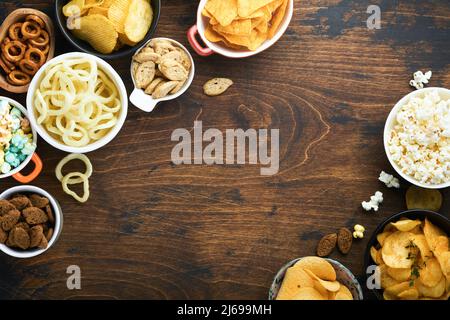 Cibo o spuntini malsani. Tutti i classici spuntini di patate con arachidi, anelli di popcorn e cipolle e pretzel salati in piatti da ciotola su vecchio sfondo di legno. Foto Stock