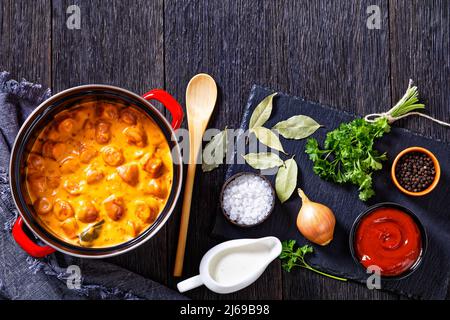 nakkikastike, salsicce finlandesi, amalgami in salsa di pomodoro cremosa in pentola rossa su tavola di legno scuro con ingredienti, vista orizzontale dall'alto, posa piatta, frè Foto Stock