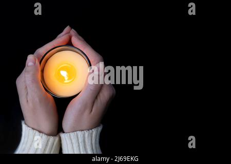 Donna che tiene con due mani una candela che brucia fiamma gialla, primo piano, su uno sfondo nero. Foto Stock