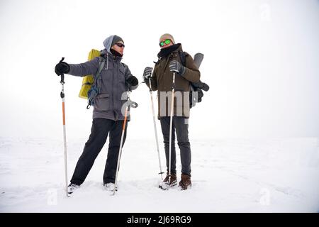 Due ragazzi camminano attraverso la neve sciolta durante una spedizione invernale. Trasportano grandi zaini, giacche calde. Tengono i bastoni da trekking nelle loro mani. Foto Stock