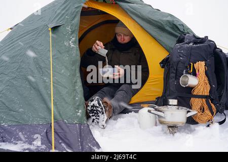 I ragazzi sono seduti in una tenda, preparando il cibo durante un'escursione invernale. Vicino alla tenda nella neve si trova la loro attrezzatura. Due uomini stanno mangiando nella tenda. C Foto Stock