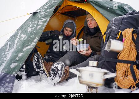 I ragazzi sono seduti in una tenda, preparando il cibo durante un'escursione invernale. Vicino alla tenda nella neve si trova la loro attrezzatura. Due uomini stanno mangiando nella tenda. C Foto Stock