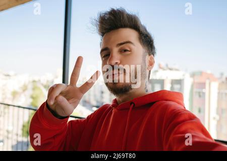 Bel giovane uomo con cappuccio rosso che prende un autoritratto all'aperto sul balcone. Ragazzo felice sorridendo alla macchina fotografica. Foto di bel giovane felice ma Foto Stock