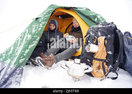 I ragazzi sono seduti in una tenda, preparando il cibo durante un'escursione invernale. Vicino alla tenda nella neve si trova la loro attrezzatura. Due uomini stanno mangiando nella tenda. C Foto Stock