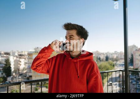 Giovane uomo con cappuccio rosso che parla con sorrisi. Uomo felice che guarda via e parla con gli amici al telefono. Foto Stock