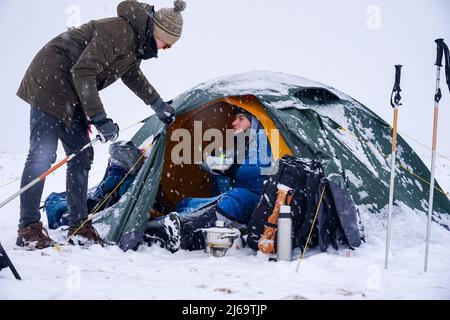 I ragazzi sono seduti in una tenda, stanno preparando il cibo durante il viaggio invernale. Vicino alla tenda nella neve è la loro attrezzatura Foto Stock