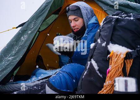 I ragazzi sono seduti in una tenda, stanno preparando il cibo durante il viaggio invernale. Vicino alla tenda nella neve è la loro attrezzatura Foto Stock