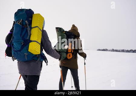 Due ragazzi camminano attraverso la neve sciolta durante una spedizione invernale. Trasportano grandi zaini, giacche calde. Tengono i bastoni da trekking nelle loro mani. Foto Stock