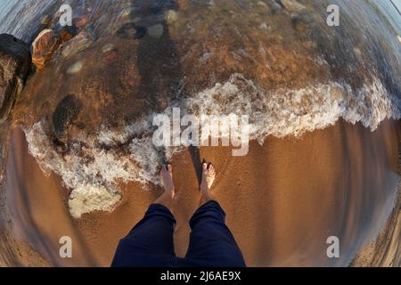 Prospettiva Fisheye in prima persona dei piedi dell'uomo in acque poco profonde con onde che si rotolano in Sandy Beach Foto Stock