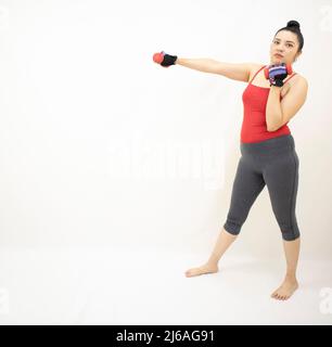 donna atletica carina che indossa abbigliamento sportivo grigio, top rosso, pratica boxe, manubri, lanciando pugni in avanti con il braccio destro, su sfondo bianco Foto Stock