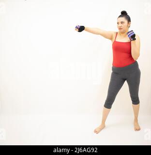 bella atletica donna che indossa abbigliamento sportivo grigio, top rosso, pratica boxe, lanciando risolutamente pugni in avanti con il braccio destro, su sfondo bianco Foto Stock