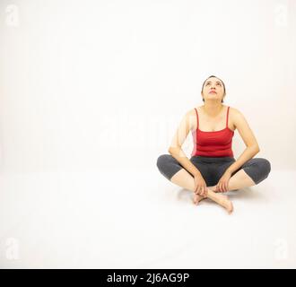 bella donna asiatica in abbigliamento sportivo grigio, blusa rossa, seduta concentrata, testa e sguardo in su, tenendo una gamba, su sfondo bianco Foto Stock