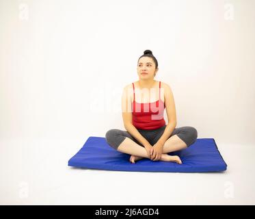bella donna in abbigliamento sportivo grigio, blusa rossa, guarda su gambe incrociate poggianti su un tappetino blu, su sfondo bianco Foto Stock