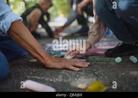 Primo piano della coppia senior con i nipoti che disegnano con i gessi sul marciapiede all'aperto nel parco. Foto Stock