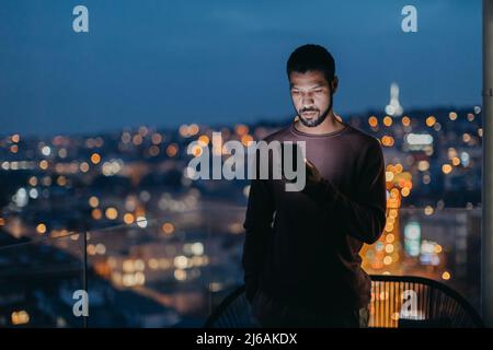 Il giovane uomo con uno smartphone sembra triste, in piedi sul balcone con vista della città di sera a casa. Foto Stock