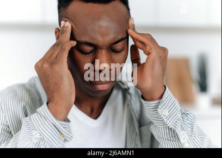 Primo piano di esausto ragazzo afroamericano in stress, si siede a casa da solo, in abiti casual, vivendo fatica, mal di testa, emicrania, templi massaggiati, chiude gli occhi, ha bisogno di riposo, medicine, trattamento Foto Stock
