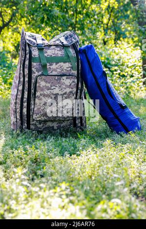 Zaino militare defocus e tenda blu o sacco a pelo. Borsa esercito su sfondo verde erba vicino albero. Zaino militare mimetizzazione. Verticale. Da a. Foto Stock