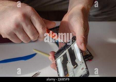 Repair of smartphones concept. A man unscrews the screws fixing the display from the board. Part of a series. Inside the room. Selective focus. Foto Stock