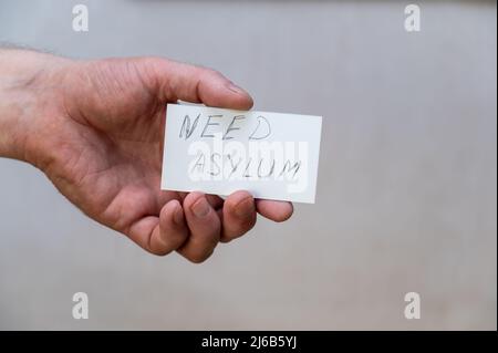 Bisogno di asilo. Parole scritte in lettere frastagliate. La mano di un uomo tiene un rettangolo di carta bianco con il testo su uno sfondo grigio. Foto Stock