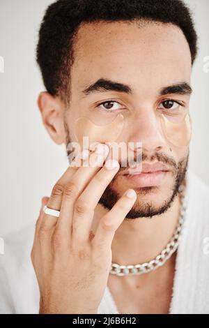 Ritratto di giovane uomo metrosessuale con accessori che applicano patch occhi sul suo viso e guardando la fotocamera Foto Stock