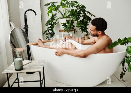 Giovane bell'uomo che rasa le gambe con un rasoio mentre prende un bagno in bagno Foto Stock
