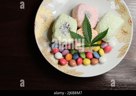 Dessert di cannabis. Delizia turca con fiocchi di cocco e confetti di caramelle colorate, vista dall'alto. Foglie verdi di cmarijuana su sfondo bianco della piastra. Layout Foto Stock