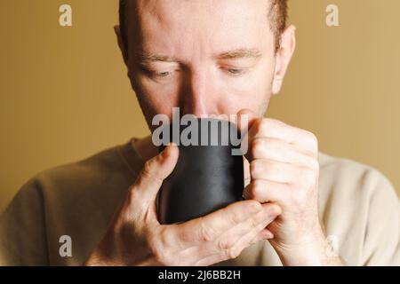 Uomo bianco non rasato che beve da una tazza. Ritratto di un uomo che beve da una tazza nera, primo piano. Foto Stock
